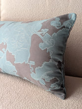 Cargar imagen en el visor de la galería, A rectangular lumbar throw pillow with a muted floral jacquard pattern in dusty blue and taupe, resting on a textured beige sofa.