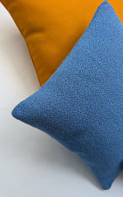 Cargar imagen en el visor de la galería, Blue textured pillow on a white surface with an orange pillow in the background